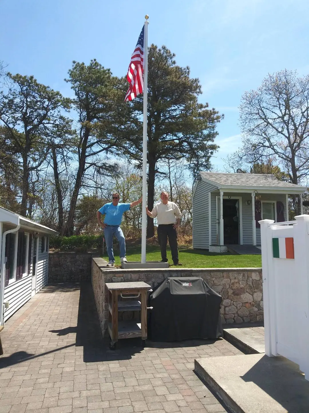 Two men stand near a raised American flag outside on a sunny day. They are on a stone patio with a small building and trees in the background.