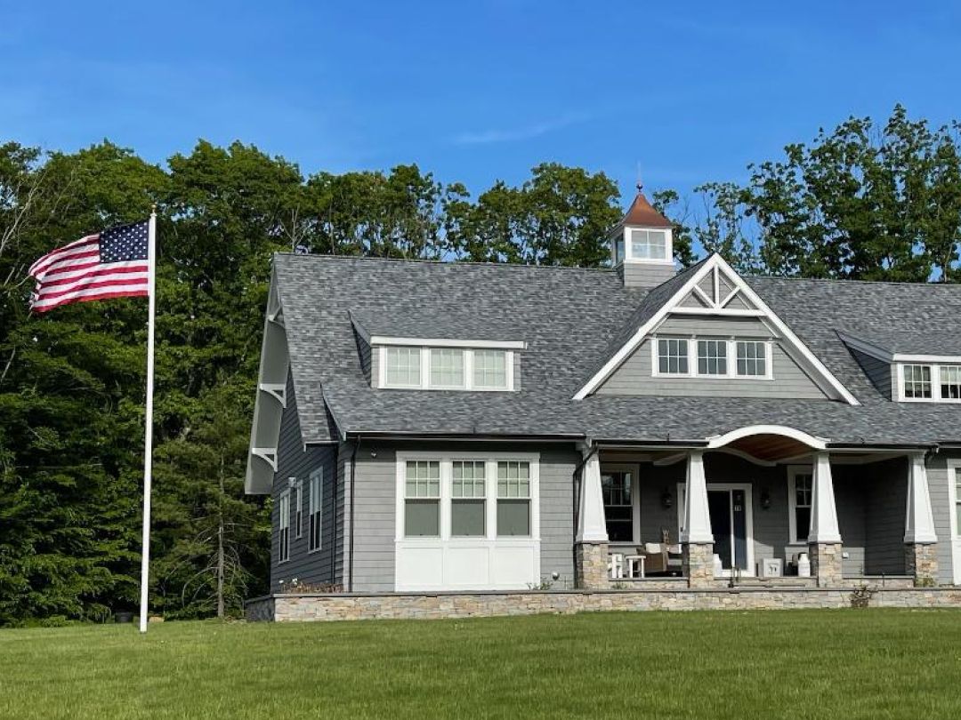 img 10 A large gray house with a steep roof and white trim, surrounded by green lawn and trees. An American flag flies on a pole beside it under a clear blue sky.