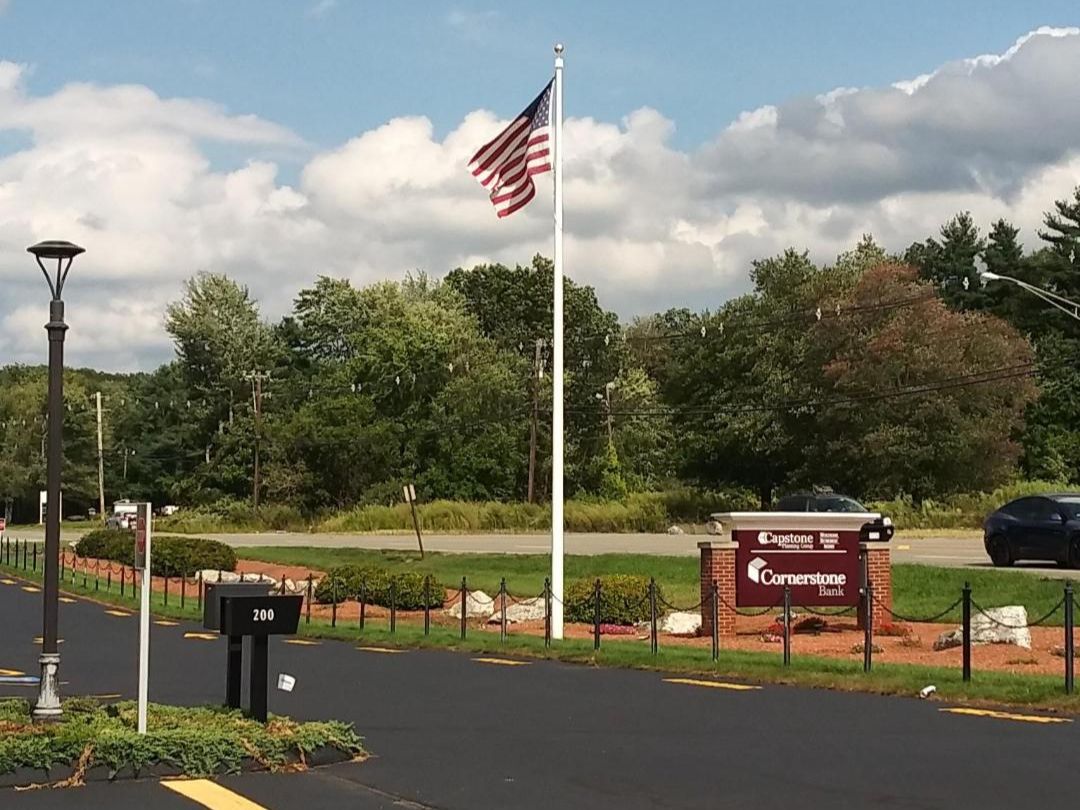 img 4 American flag on a pole near a bank sign, surrounded by greenery and a parking area under a partly cloudy sky.