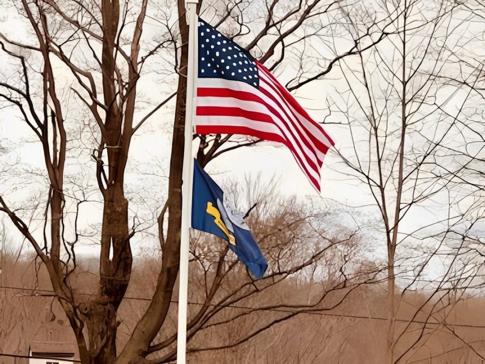 img-6 (1) Two flags on flagpoles: a U.S. flag above and a blue flag below, with bare trees in the background.
