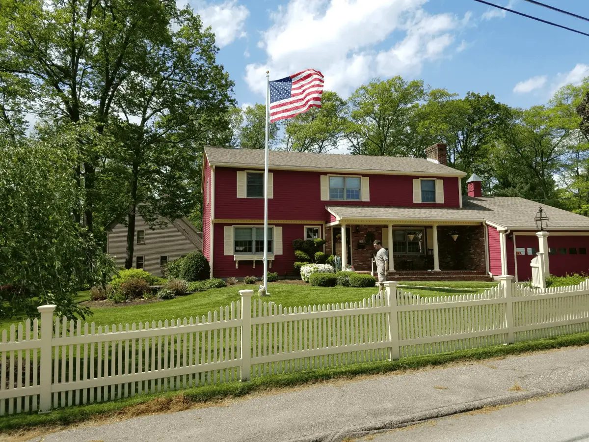 A red two-story house with a white picket fence and an American flag.